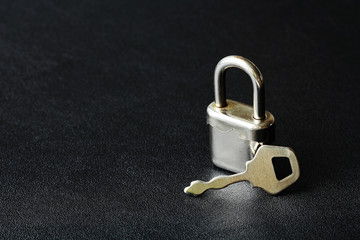 Small silver closed lock with a key on a black leather background. Copy space