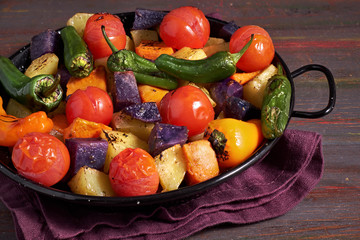 Rustic, oven baked vegetables in baking dish. Seasonal vegetarian vegan meal on dark wooden background with linen towel.