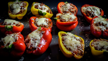 Red and yellow bell peppers cut in half, filled with minced beef meat, topped with cheese and baked, on dark background
