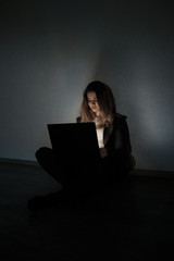 Sad teenage girl sitting near laptop in dark room. She is a victim of online bullying Stalker...