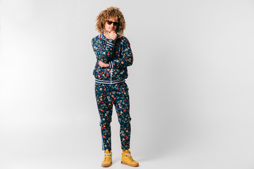 Adult shylish man in fashionable tracksuit with flowers pattern, yellow sneakers posing on white background. Funky man stroking shaved beard. Awesome confident funky male in sunglasses portrait.