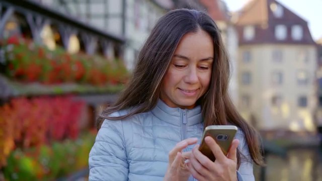Close-up shot of a smiling brunnete woman in blue jacket going through her photos on smartphone outdoor.