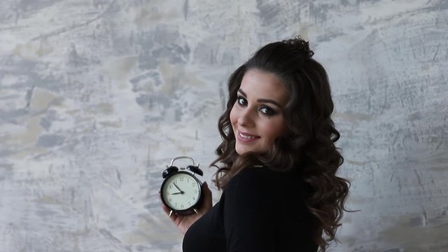 Beautiful pregnant woman in black bodysuit. She hold the clock in hands symbolic hinting at the remanining time befour the baby in born. Woman put her hand on belly. close up
