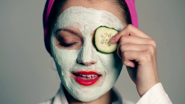 Beautiful woman with mud mask cucumber slices. Beautiful girl holding a slice of cucumber in front of her face. Photo of girl in white bathrobe and with mask of clay on face.