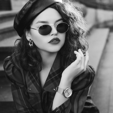 Monochrome close up fashion portrait of young beautiful fashionable girl wearing trendy sunglasses, wrist watch, earrings, leather beret, checkered dress, posing in street, sitting on stairs