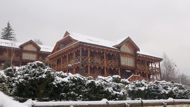 Wooden cottages in mountain village during heavy snowfall with coniferous forest on background. Snow falling on log chalets at ski resort with evergreen landscape design. Cold frosty winter day.