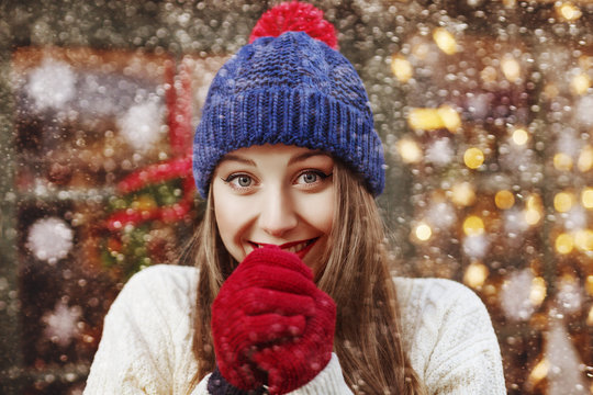 Street portrait of young beautiful happy smiling girl warming her hands with her mouth. Lady wearing blue beanie hat, red gloves, white sweater. Christmas, winter holidays concept. Snowfall. Close up