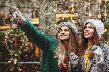 Happy smiling surprised girl pointing up to her friend. Models looking up, wearing stylish winter clothes. Snowfall, festive Christmas background.
