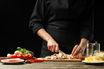 The chef cuts champignon mushrooms with vegetables. For the preparation of pizza, mushroom sauce, salad. A delicious food concept, cooking. On a black background for design or lettering text
