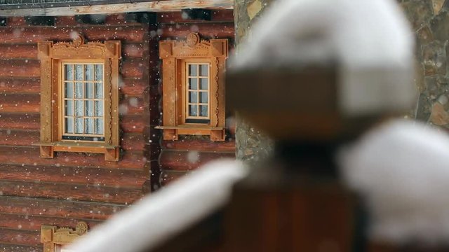Close view of cottage with classic windows in mountain village during heavy snowfall with russian style carved wooden facade. Snow falling on log chalets at ski resort. Cold frosty winter day.