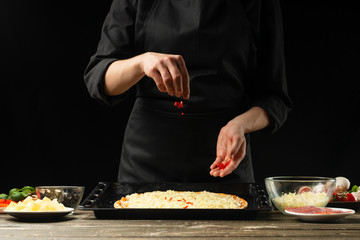 The chef sprinkles pizza with chilli peppers. A concept of delicious food and healthy food. On a black background for design or lettering text.