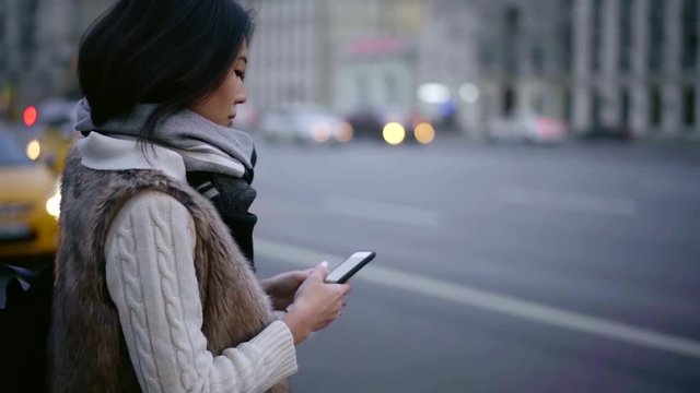 Brunette firl standing by the road with a smartphone and ordering a taxi.