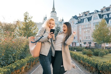 Obraz premium Two young women having fun, looking at the smartphone laughing, sunny autumn day, city background, golden hour
