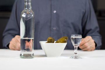 Butelka wódki, ogórki kiszone, kieliszek z wódką stoją na stole przykrytym białym obrusem. W tle postać mężczyzny siedzącego za stołem. 