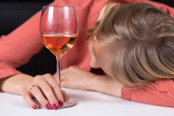 Kobieta śpi przy stole z głową opartą o rękę. Kieliszek z winem trzyma w ręce.