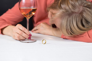 Kobieta śpi przy stole z głową opartą o rękę. Kieliszek z winem trzyma w ręce. Złota ślubna obrączka leży na stole przed nią. Kłopoty małżeńskie.