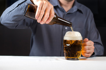 Mężczyzna siedzi przy stole i nalewa jasne piwo z butelki do kufla. 