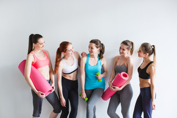 Group of young sporty woman with yoga mats standing at white wall.