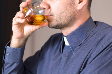 Ksiądz w koloratce i szarej koszuli pije whisky z lodem ze szklanki.