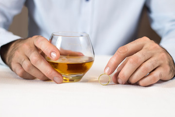 Kłopoty małżeńskie. Dłonie mężczyzny siedzącego przy białym stole trzymają szklankę z whisky. Druga ręka trzyma obrączkę ślubną. - obrazy, fototapety, plakaty