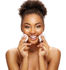 Charming young girl with flawless skin is holding cotton pads near face. Photo of smiling african american girl on white background. Skin care and beauty concept