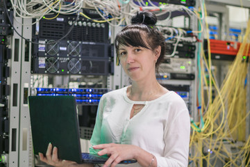 Girl with a laptop is in the server room. Programmer works on the computer in the data center. Woman is on the background of powerful servers. Concept of technical support and computer administration