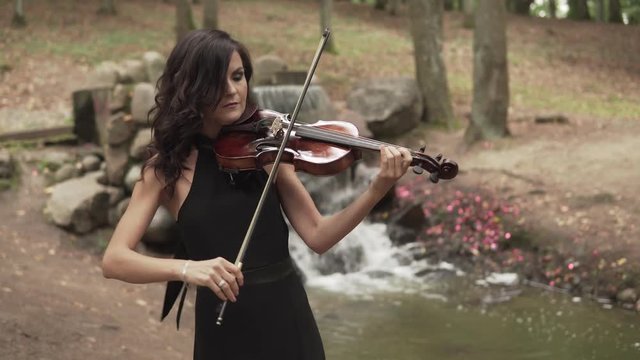 Beautiful girl in black dress playing violin in forest with waterfall at background. Brunette violinist plays with inspiration