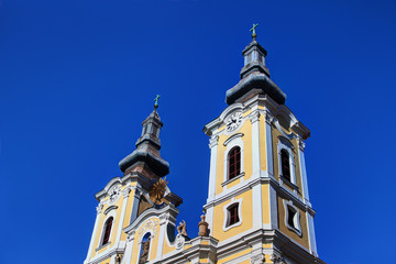 Fototapeta na wymiar Low-angle view of two yellow and white clock towers of sunlit Baroque Minorite church against clear blue sky, downtown Heroes' Square in Hungarian city Miskolc, Borsod county Central / Eastern Europe