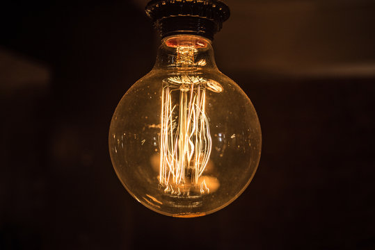 Large long electric incandescent filament in a light bulb glows and reflects warm light.