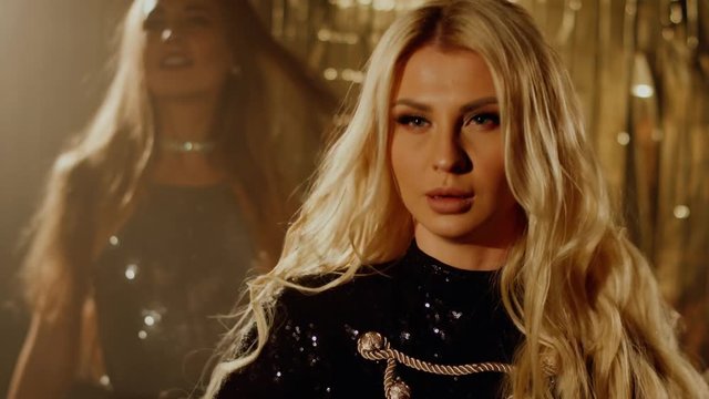 Alluring blonde woman and her female friend are resting on a dancefloor in clubhouse. They are dancing rhythmically by music, close-up of faces. Shoot with RED RAVEN camera. 