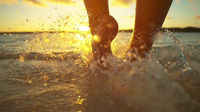 SLOW MOTION, CLOSE UP, LOW ANGLE: Glassy drops of water splash around the unrecognizable girl's feet as she goes on a relaxing walk along the sandy beach on a picturesque sunny evening in Cook Islands