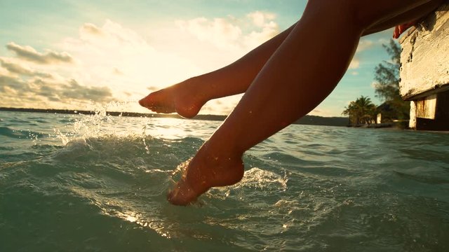 SLOW MOTION, LENS FLARE, CLOSE UP: Playful female tourist on relaxing summer holiday on paradise island splashing water with her bare feet on a sunny morning. Carefree girl kicking the ocean water.