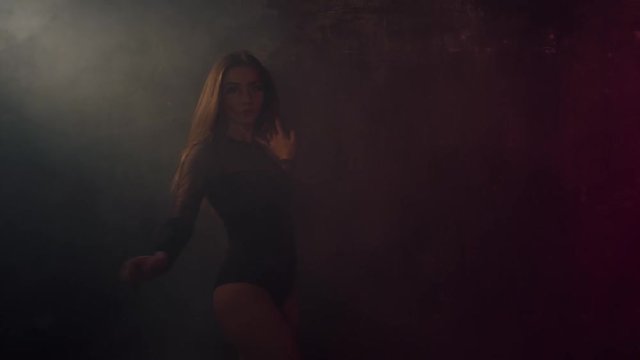 Charming girl is dancing sensually in darkness in dance floor. She is whirling, leaning on wall and lowering in smoke. Shoot with RED RAVEN camera. 