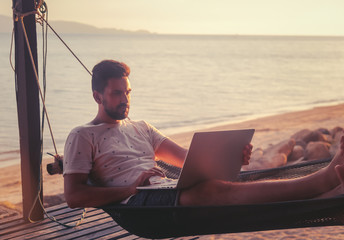 Young attractive guy a man with a beard in a hammock with a laptop on the background of the sea and...