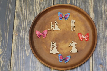 Romantic composition on ceramic dish. Stylized wooden figures of lovers and colorful ornamental satin butterflies. Dark wooden background.