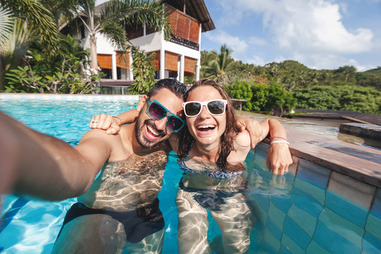 Attractive young couple in love is having fun in the outdoor pool, taking a selfie on a mobile phone; luxury luxury vacation, real estate in the tropics, honeymoon - Powered by Adobe