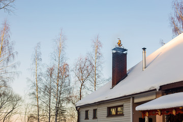 Part of snow-covered roof of house with pipe from which there is smoke on background of high birches and endless blue sky