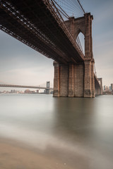 Obraz premium Widok na Brooklyn Bridge od wschodniej rzeki piaszczystej plaży o zachodzie słońca z długim czasem ekspozycji