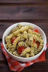 White bowl of fusilli with green basil pesto and cherry tomatoes, rustic wooden background, vertical shot