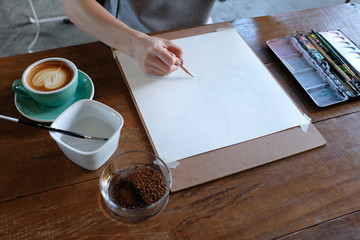 A woman's hand holding a brush painting a coffee cup on white paper with coffee paint on wooden table with props