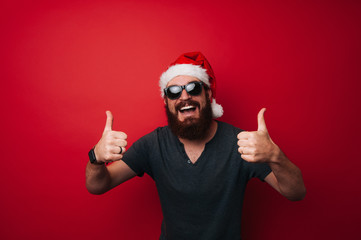Smiling man with beard wearing santa claus hat showing thumbs up gesture over red background