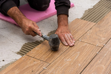 Worker placing ceramic floor tiles on adhesive surface, leveling with rubber hammer.