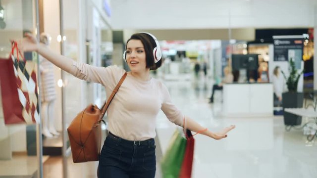 Happy female shopaholic is having fun in shopping mall listening to music in headphones, dancing with bright bags and laughing pointing at goods in shop windows.