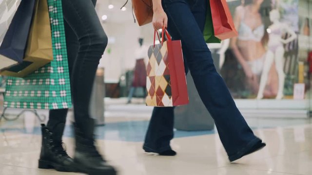 Low angle shot of slim girls wearing jeans walking in shopping mall together carrying colorful paperbags after day of purchases. Shopaholics, youth and stores concept.