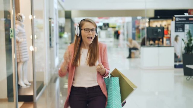 Happy woman is enjoying music in headphones, dancing and singing in shopping mall hall holding paper bags. Girl is looking at clothing in shop windows and having fun.