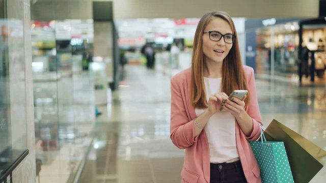 Cute girl with fair hair is using smartphone and smiling while walking in shopping mall with paper bags. Internet, modern technology and youth lifestyle concept.