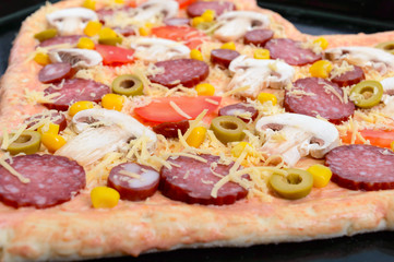 Delicious pizza in a heart shape, ready for baking. Pizza with mushrooms, salami, pepperoni, olives, corn on a baking tray.
