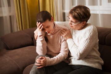 senior mother consoles, comforting sad adult grown daughter sitting at sofa at home. support in two...