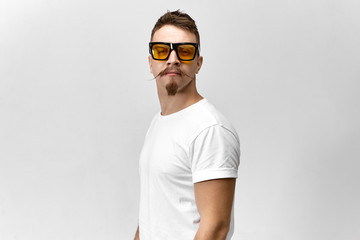 Youth, fashion and fun. Picture of attractive funny young Caucasian male with goatee beard and curled mustache looking at camera wearing stylish glasses with yellow tinted lenses for better vision