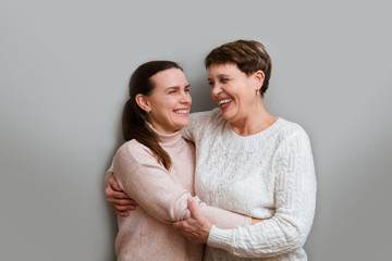 Happy senior lady mother and adult daughter embracing looking at camera on a gray background